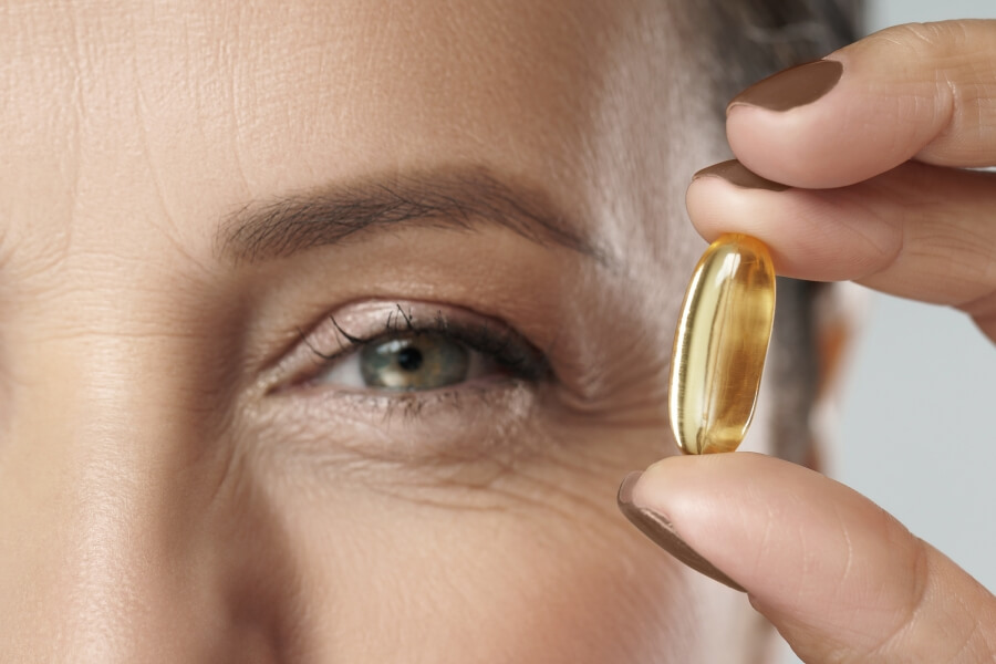 Detail ženy, která drží kapsli s omega-3 mastnými kyselinami u oka – vliv omega 3 na zrak a zdravé stárnutí