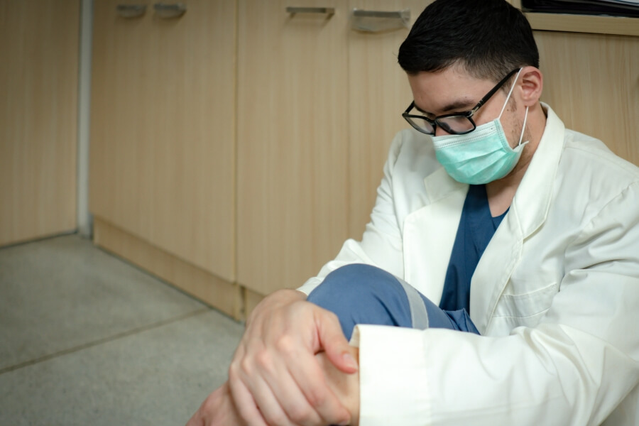 Unavený zdravotník sedící na zemi v ordinaci, držící se za kolena, vyjadřující hluboké pracovní přetížení a příznaky syndromu vyhoření u zdravotníků