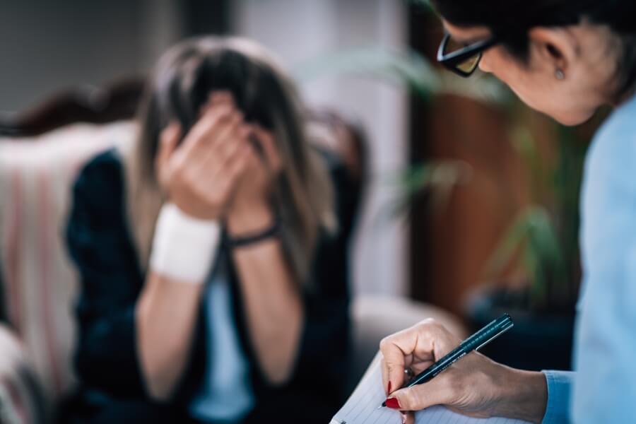 Žena zakrývající si obličej rukama při rozhovoru s terapeutkou, ilustrující silné emoční vyčerpání, psychickou krizi a potřebu odborné pomoci při syndromu vyhoření