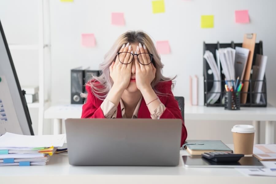 Mladá žena v červeném saku sedící u notebooku a zakrývající si obličej rukama, typický obraz emočního vyčerpání a pracovní frustrace při syndromu vyhoření